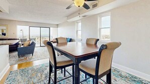 A spacious room with a dining table, chairs, a ceiling fan, and large windows.