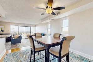 A spacious room with a dining table, chairs, a ceiling fan, and large windows. - Capri Condo: Bay View balcony, WiFi, AC, W/D & shared pool/gym/tennis/sauna (Ocean City)