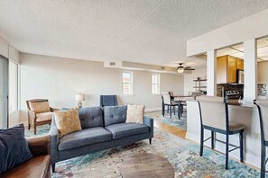 A living room with a sofa, chairs, a coffee table, and a kitchen area with a dining table and chairs. - Capri Condo: Bay View balcony, WiFi, AC, W/D & shared pool/gym/tennis/sauna (Ocean City)