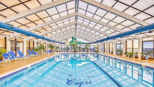 An indoor swimming pool with a glass ceiling, blue lounge chairs, and potted plants.