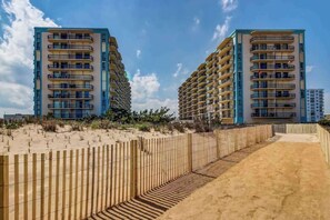 Un hôtel situé en bord de mer, doté de nombreux balcons, d’une promenade de sable et d’une clôture en bois.