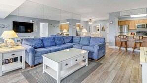 A modern living room with a blue sofa, a glass coffee table, and a kitchen area with wooden cabinets and a microwave. - Braemar Towers 1006 (Ocean City)