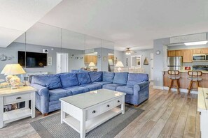 Un salon moderne avec un canapé bleu, une table basse en verre et un coin cuisine doté de meubles en bois et d’un four à micro-ondes.