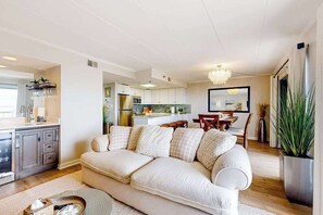 A modern living room with a beige sofa, a dining area with a wooden table, and a kitchen in the background. - Bay Princess 403 - Just Breathe (Ocean City)