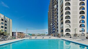A swimming pool with lounge chairs, surrounded by high-rise buildings. - Third-floor condo in oceanfront building with balcony, pool & beach access (Ocean City)