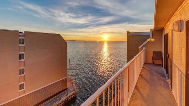 Un balcone con vista sul tramonto sull'acqua, un edificio a tetto piatto e una ringhiera.