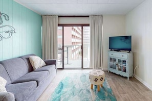 A living room with a sofa, ottoman, television, cabinet, and a view through a glass door to an outdoor balcony. - Ocean-view condo with balcony & pool access - steps from the beach (Ocean City)