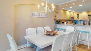 A dining area with a white table and chairs, a chandelier, and a kitchen in the background. - Adagio 407 (Ocean City)