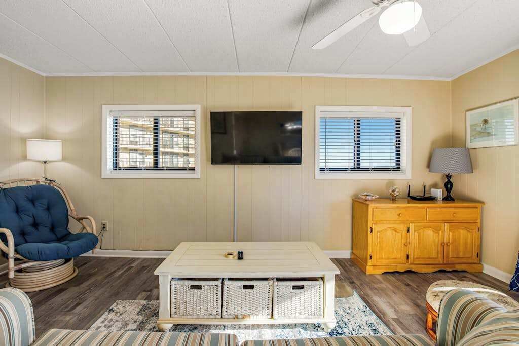 A living room with a television, wicker chair, striped sofa, wooden cabinet, and a ceiling fan.