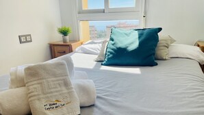 A neatly made bed with white linens and a teal pillow, a small wooden nightstand with a plant, and a window with a view of buildings. - Apartamento Faro 77 by At Home Costa del Sol (Torrox)