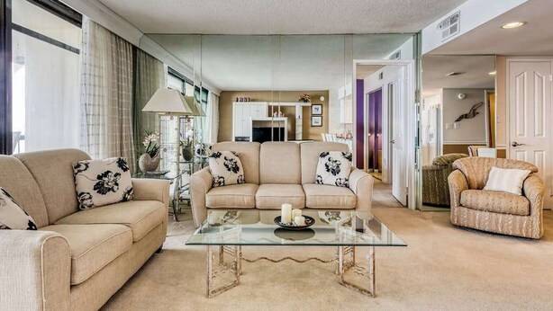 A living room with a glass coffee table, beige sofas with floral cushions, and a view into the kitchen. - Irene 1106 (Ocean City)