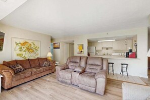 A living room with a sofa, a dining area with a kitchen in the background, and a painting on the wall. - Fourth-Floor Condo near the Beach w/Private Balcony, Ocean View, A/C, Free WiFi (Ocean City)