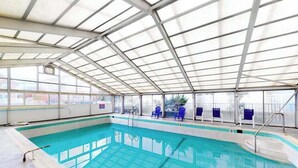 An indoor swimming pool with clear glass walls, blue chairs, and a metal roof structure. - Decatur House 203 (Ocean City)