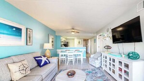 A living room with a sofa, a coffee table, a TV, and a kitchen in the background. - Colorful, ocean-view condo with scenic balcony, 3 pools, washer/dryer & AC (Ocean City)