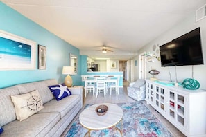 A living room with a sofa, a coffee table, a TV, and a kitchen in the background. - Colorful, ocean-view condo with scenic balcony, 3 pools, washer/dryer & AC (Ocean City)