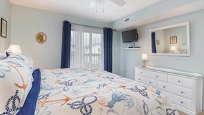 Ein Schlafzimmer mit einem Bett, einem Deckenventilator, einem Fernseher, einer Kommode und einem Fenster mit Vorhängen.