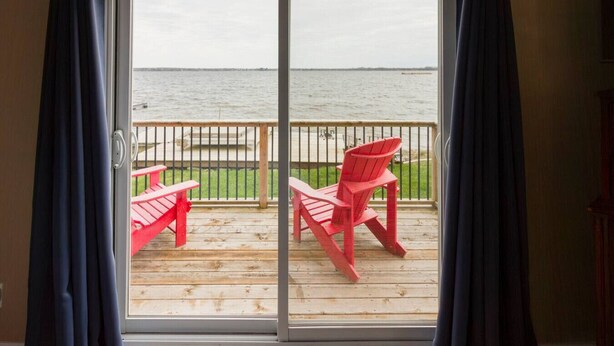 Een kamer met een schuifdeur die uitkomt op een houten terras met twee rode Adirondack-stoelen en uitzicht op het water.