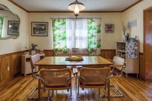Een eetkamer met een houten tafel, vier rieten stoelen, een kroonluchter, een koelkast en een muur met ingelijste foto’s en een bord.'