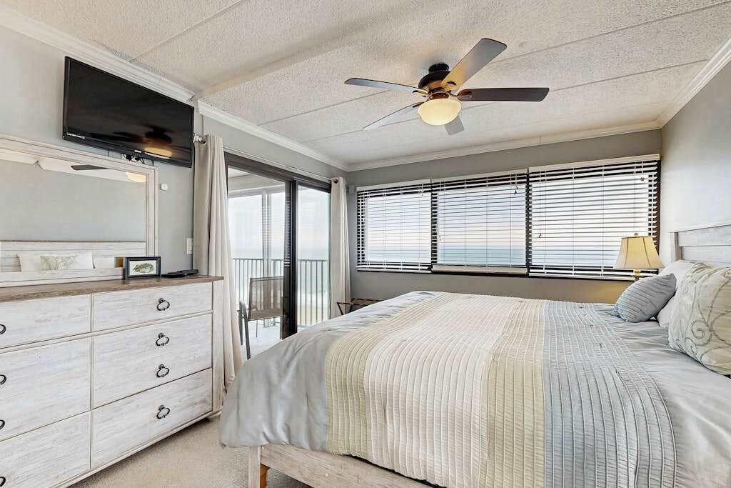 A bedroom with a bed, a dresser, a ceiling fan, a TV, and a window with blinds.