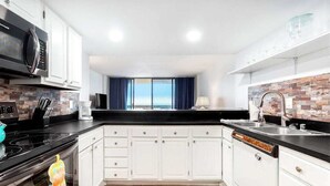 Une cuisine moderne avec des meubles blancs, un plan de travail noir, un four à micro-ondes, une cuisinière et une vue sur la mer grâce à de grandes fenêtres.