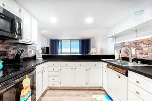 A modern kitchen with white cabinets, a black countertop, a microwave, a stove, and a view of the sea through large windows. - Oceanfront Getaway w/ Private Balcony, Free WiFi, Beach Access & Shared Pool (Ocean City)