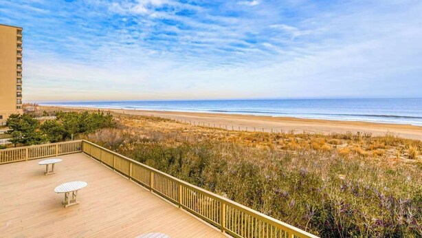 Une terrasse en bois avec deux tables, donnant sur une plage bordée de dunes et sous un ciel dégagé.