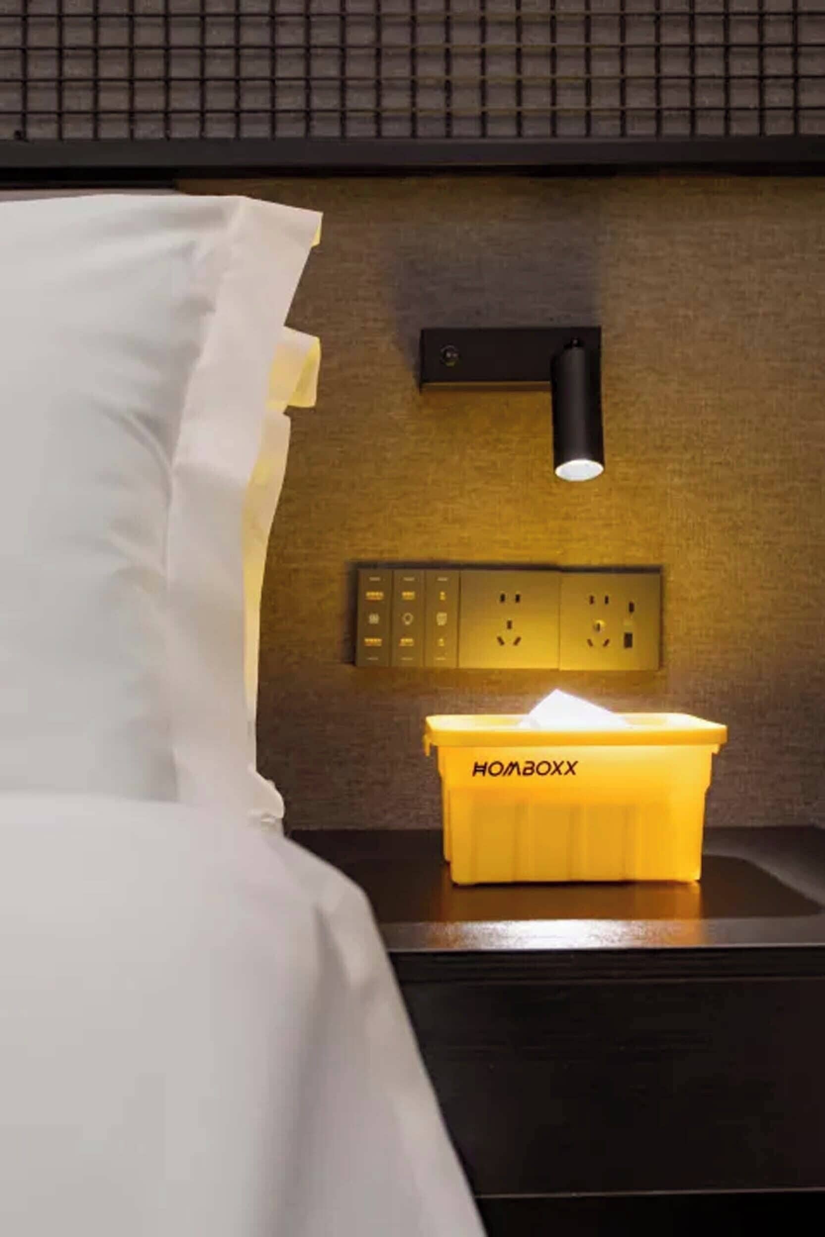 A hotel room with a bed, a nightstand with a lamp, a power outlet, and a trash bin labeled 'HOBOX.'