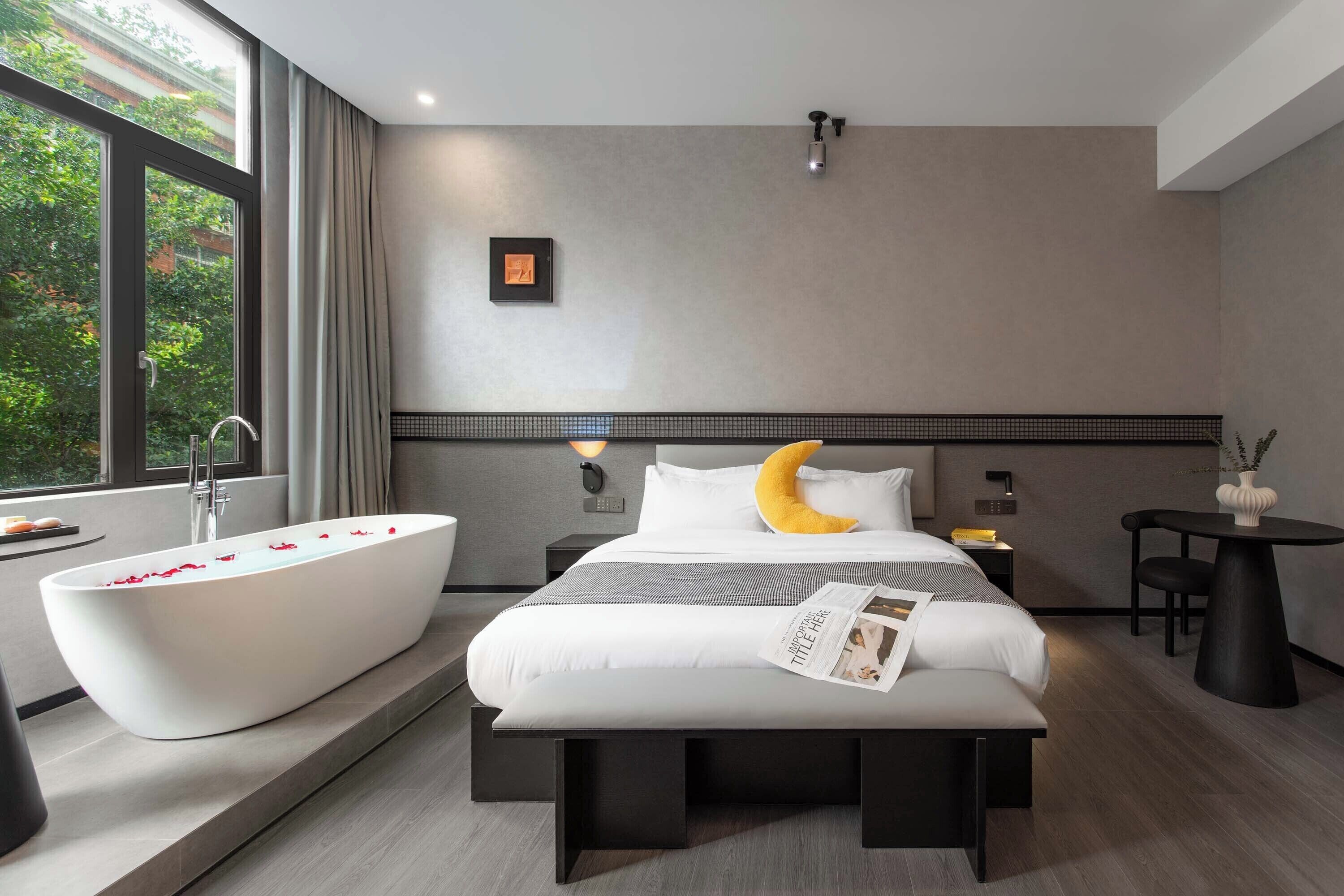 A modern hotel room with a large bathtub, a bed with a unique crescent-shaped pillow, a bedside table, and a view of greenery outside.