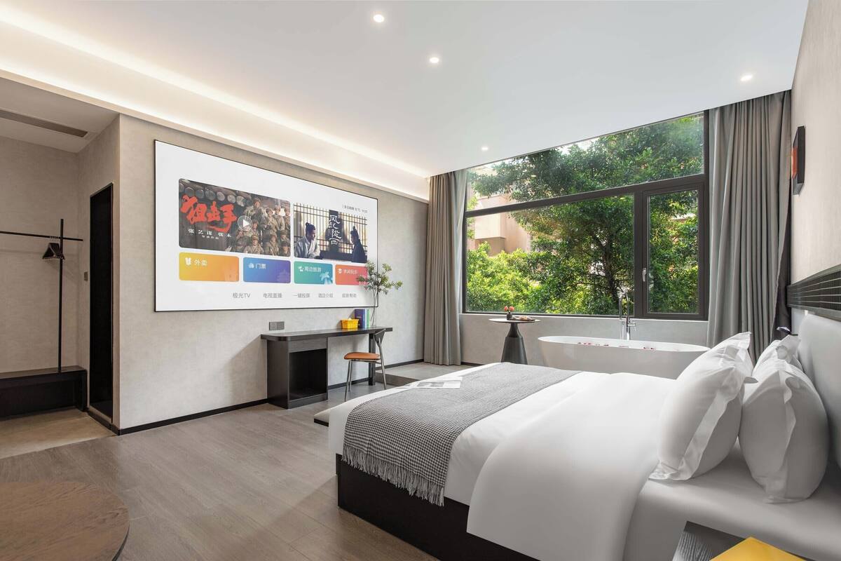 Una habitación de hotel moderna con una cama grande, un escritorio, una silla, un televisor montado en la pared y una ventana con vista a zonas verdes.