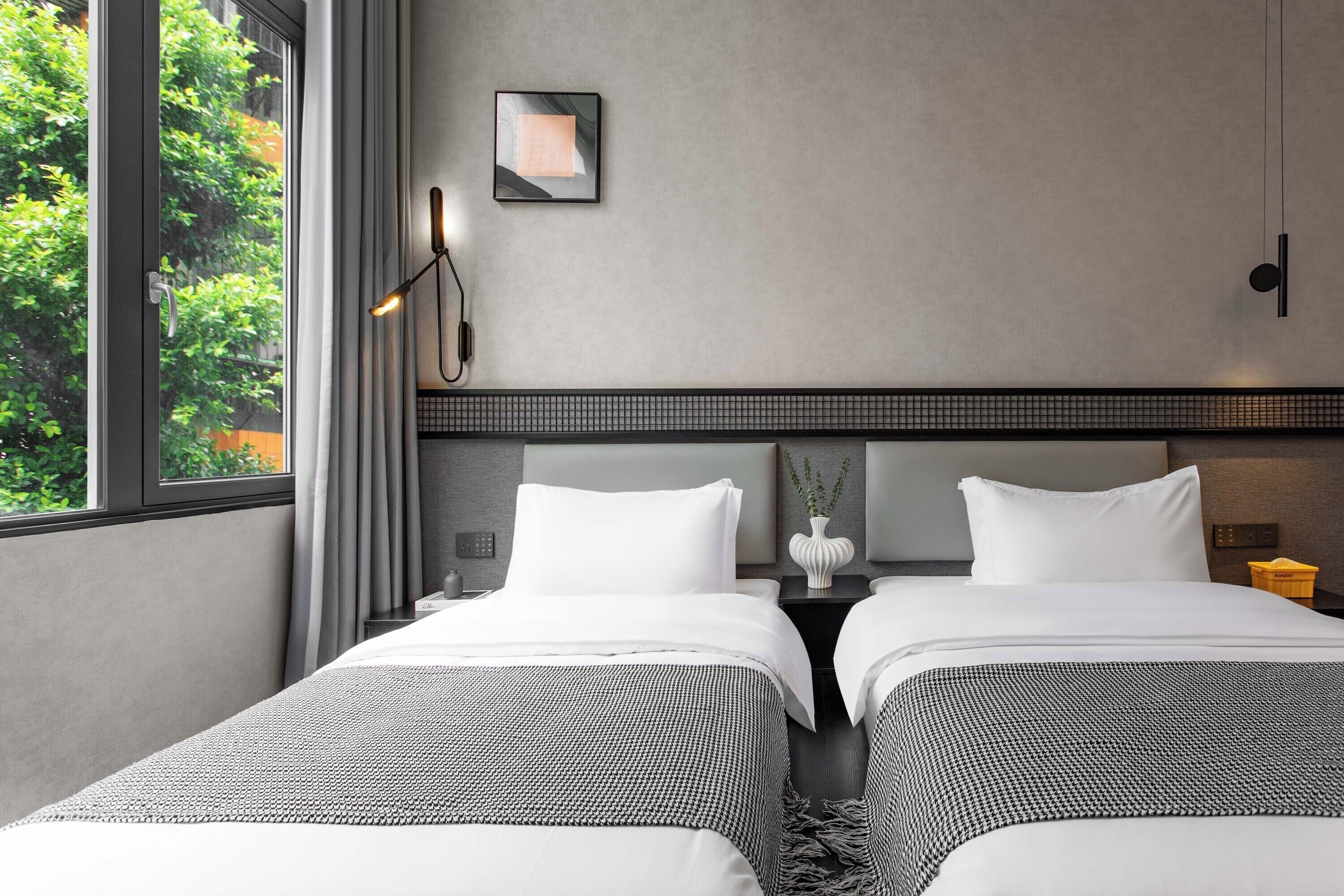 Habitación de hotel con dos camas, vistas a la vegetación, una lámpara en la pared y una mesita con un florero.