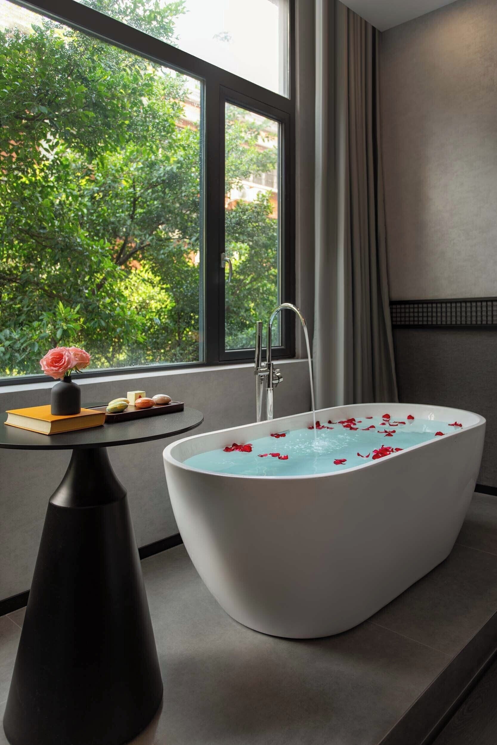Un baño moderno con una bañera independiente, una mesita con un jarrón de flores y vistas a la vegetación exterior.