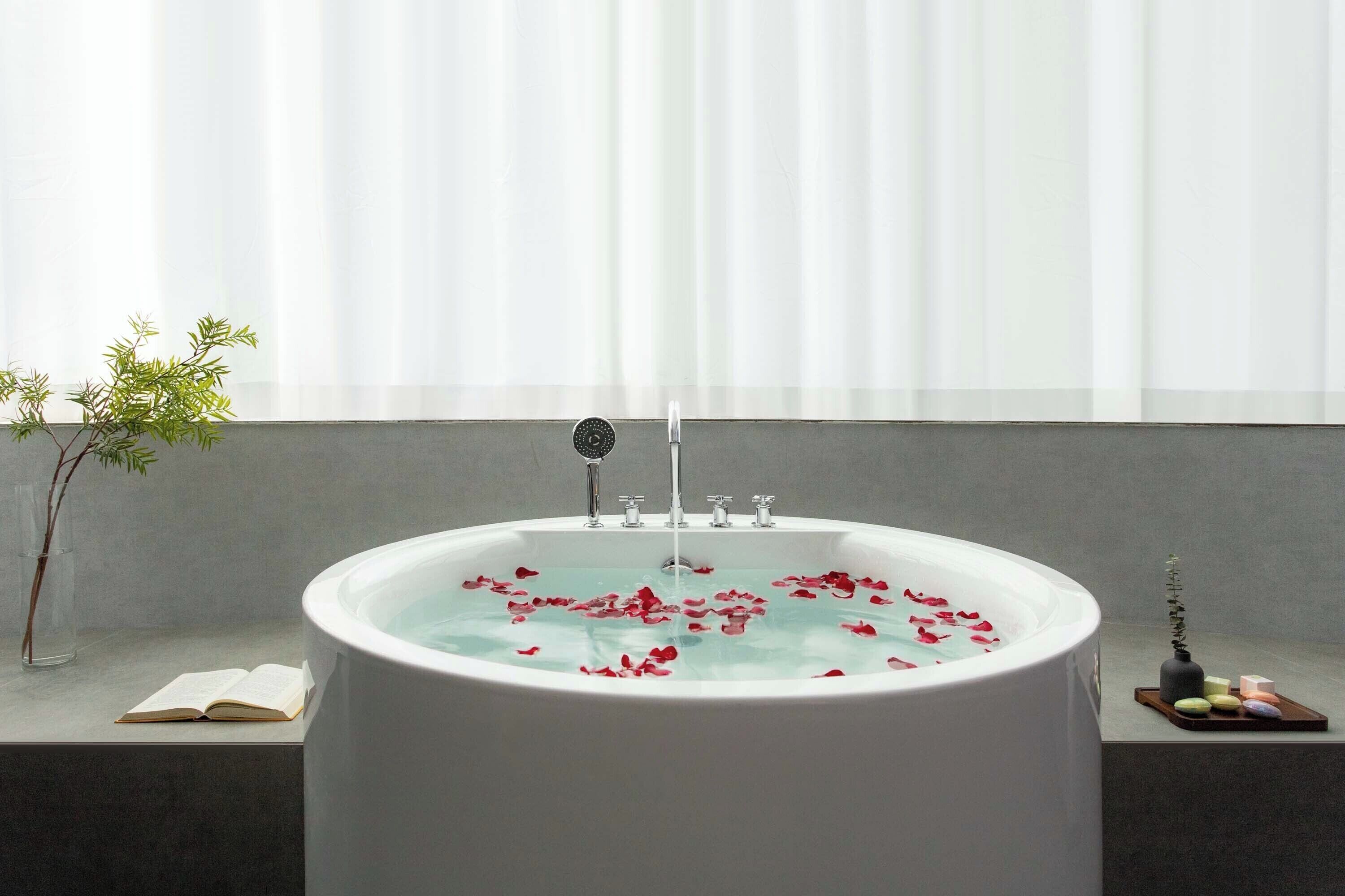 Une baignoire remplie de pétales de rose, un livre et une petite plante posée sur le rebord d’une fenêtre.