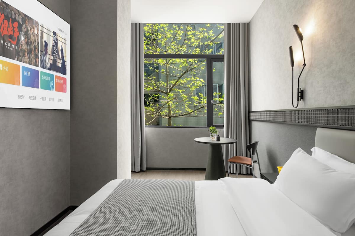 Una habitación de hotel moderna con una cama, una mesita, una silla, una lámpara de pared y un ventanal con vistas a árboles.
