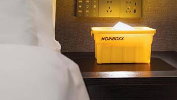 Habitación de hotel con cama, mesita de noche, lámpara de pared y un cubo de basura amarillo etiquetado 'HOMBOXX'.