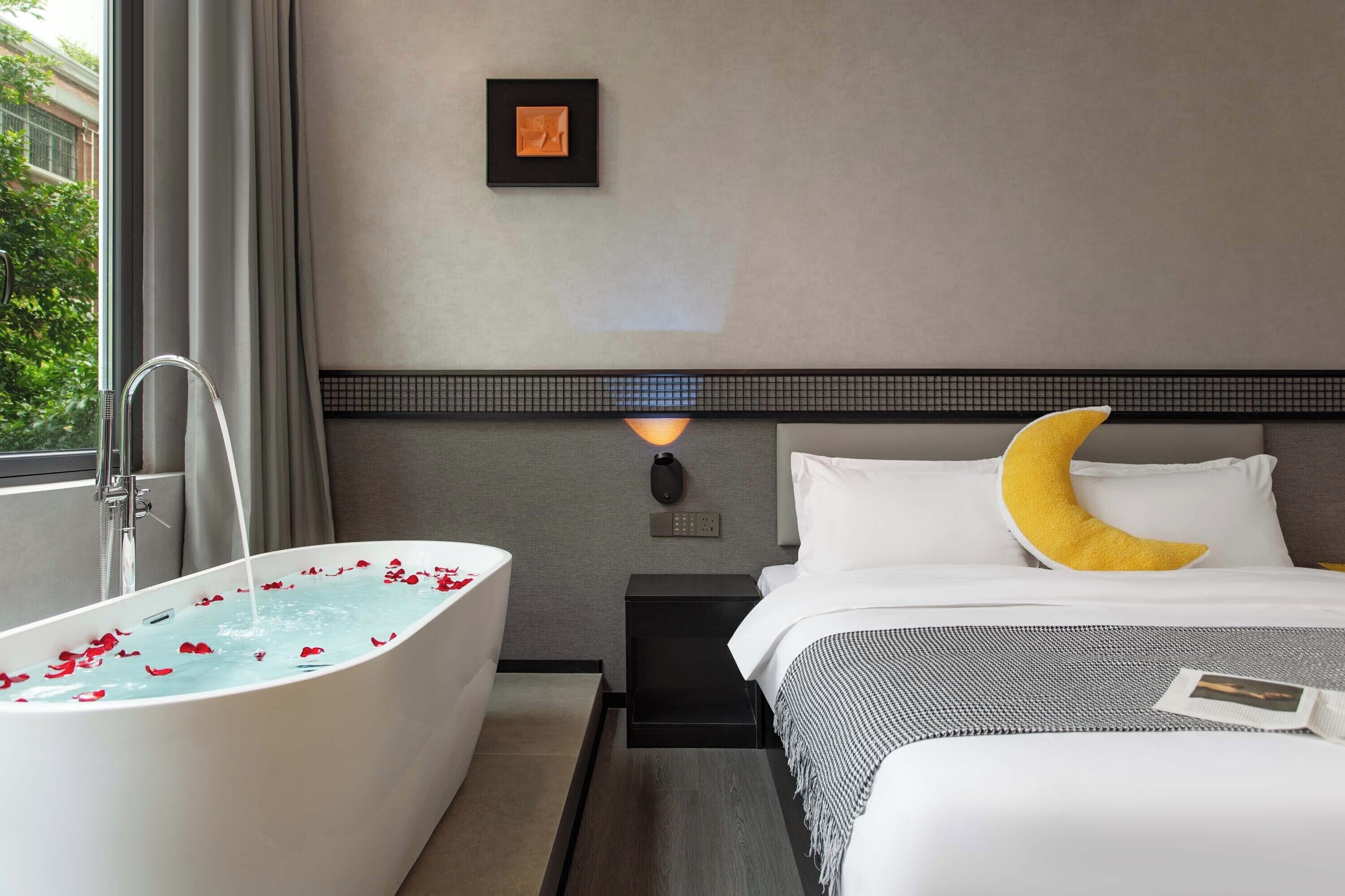 Una habitación de hotel con bañera, una cama con almohada de medialuna, una mesita de noche con lámpara y un cuadro en la pared.