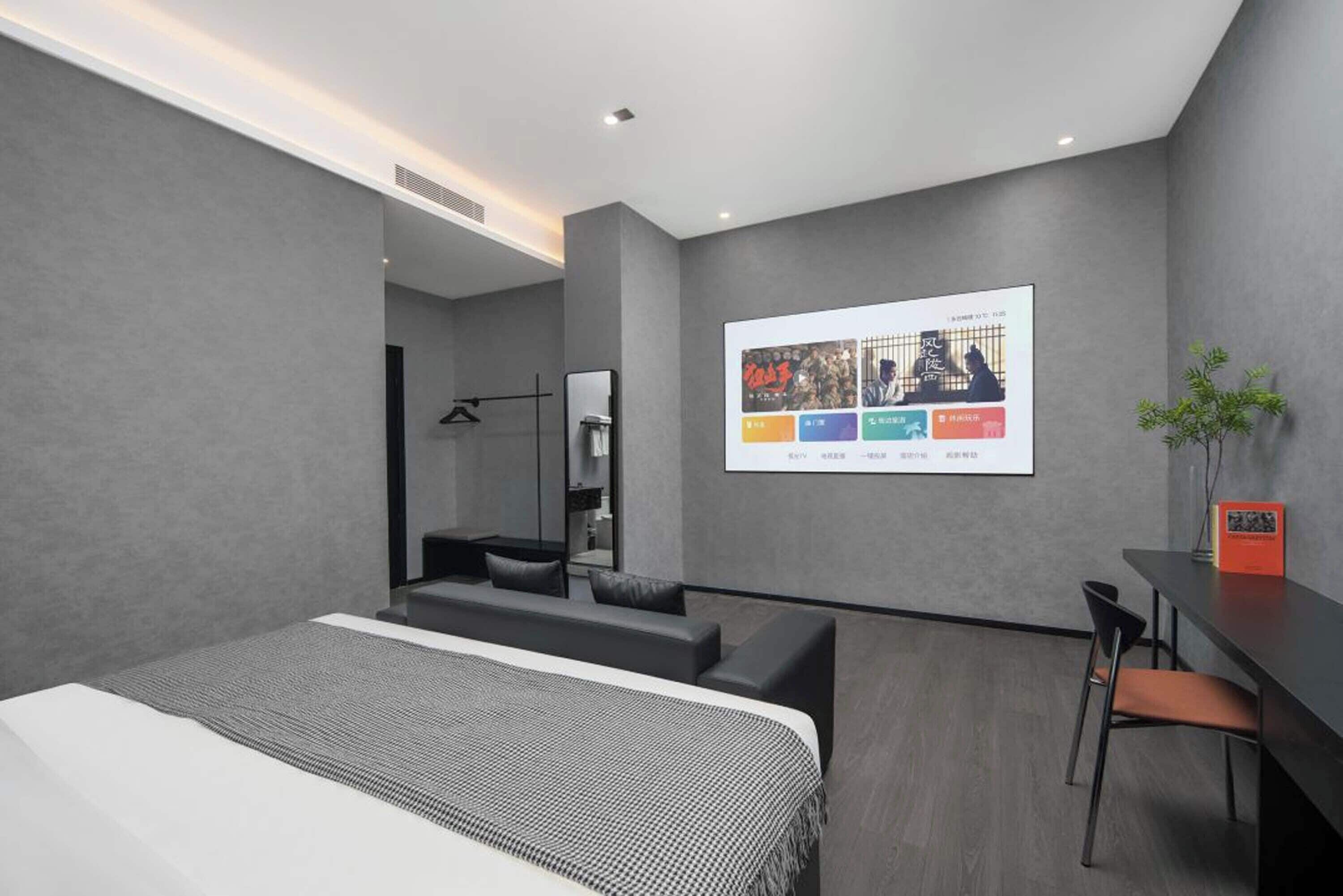 Ett modernt hotellrum med en säng, ett skrivbord, en stol, en liten sittgrupp och en väggmonterad skärm som visar en meny.