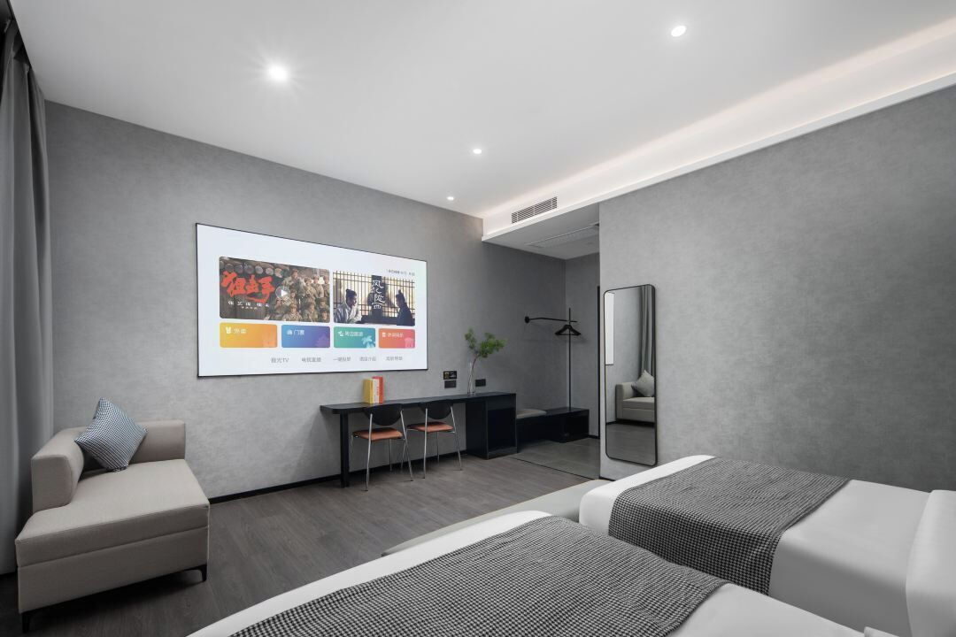 A modern hotel room with a bed, a sofa, a desk, and a large screen displaying a presentation.