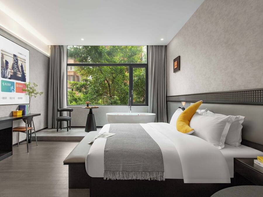 Una habitación de hotel moderna con una cama grande, un escritorio, una silla, una mesita y un ventanal con vistas a árboles.