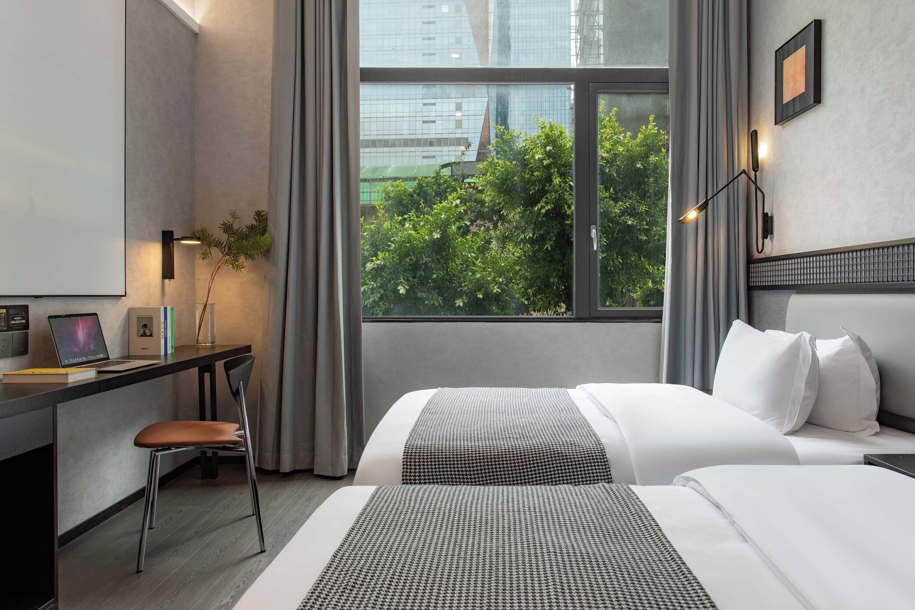 A hotel room with a desk, two beds, a chair, a window with a view of greenery, and a wall-mounted light fixture.