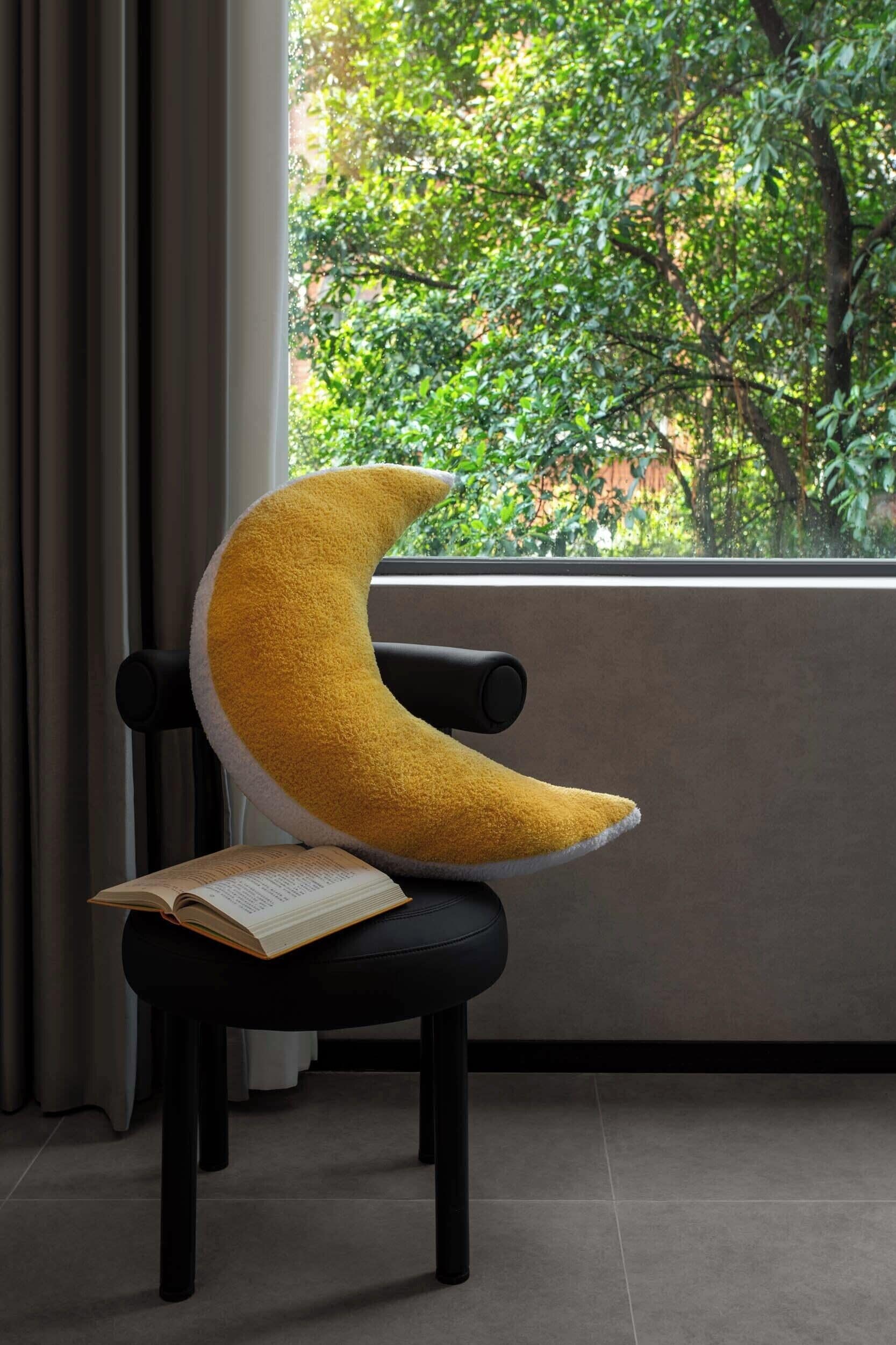 Una silla con un cojín en forma de media luna y un libro abierto encima, ubicada cerca de una ventana con vistas al verdor.