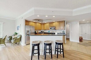 A modern kitchen with wooden cabinets, a stainless steel refrigerator, a microwave, and a bar area with stools. - 4100 #201 (Ocean City)