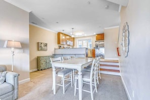 A dining area with a white table and chairs, a kitchen in the background, and a staircase leading to another level.