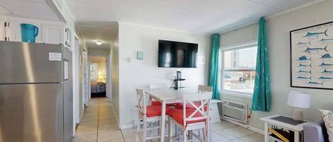 A compact kitchen with a refrigerator, a dining area with a table and chairs, a TV mounted on the wall, and a window with curtains.