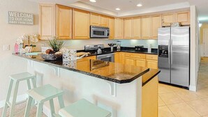 A modern kitchen with wooden cabinets, stainless steel appliances, a central island with bar stools, and a welcome sign. - Midtown Condo with Balcony, Gas Fireplace, Washer/Dryer, WiFi & Shared Pool (Ocean City)