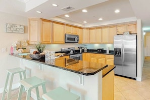 A modern kitchen with wooden cabinets, stainless steel appliances, a central island with bar stools, and a welcome sign. - Midtown Condo with Balcony, Gas Fireplace, Washer/Dryer, WiFi & Shared Pool (Ocean City)