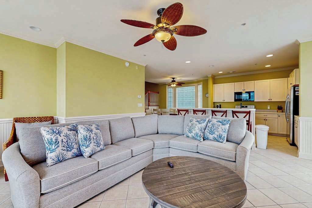 A living room with a sectional sofa, a round coffee table, a ceiling fan, and a kitchen in the background.