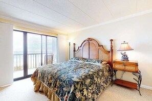 A bedroom with a large bed, a nightstand, a lamp, and a view of the outdoors. - Oceanfront Condo with Balcony, Central AC, Washer/Dryer, WiFi & Shared Pool (Ocean City)