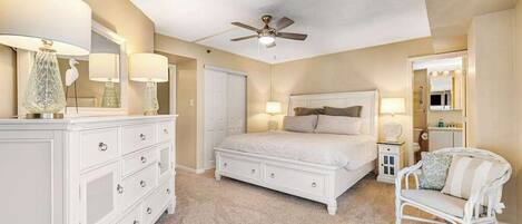 A bedroom with a bed, dresser, chair, ceiling fan, and a door leading to another room.