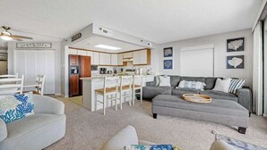 Ein modernes Wohnzimmer mit grauem Sofa, einem weißen Sessel und einem Essbereich, im Hintergrund befindet sich eine Küche.