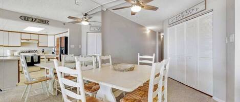 A dining area with a white table and chairs, ceiling fans, and a kitchen in the background.
